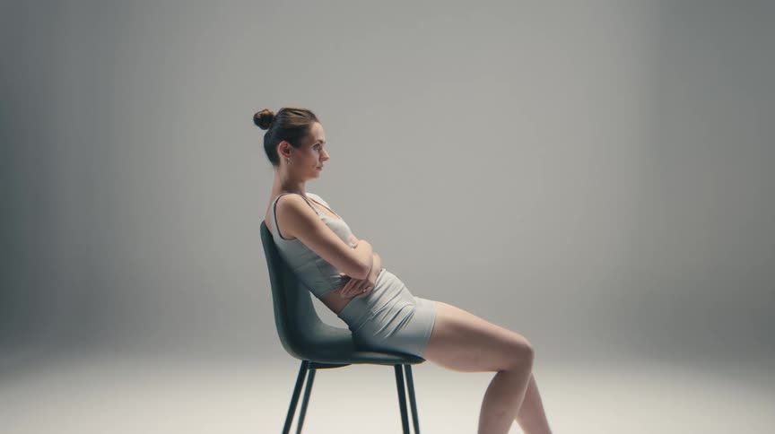 An unsmiling woman in a grey sports bra and shorts is sitting with her arms crossed against a simple grey background