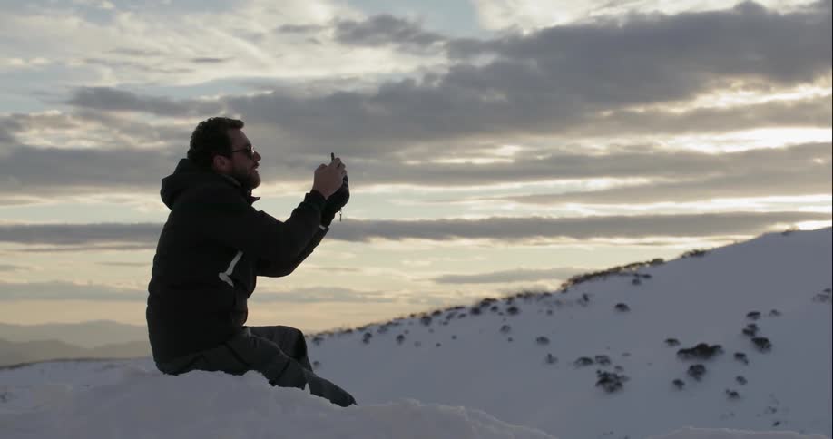A man sits in the snowy mountains and takes a picture with his phone
