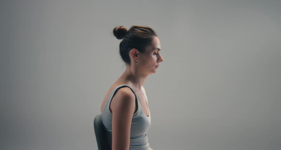 A woman with slouched posture sits in profile against a plain gray background