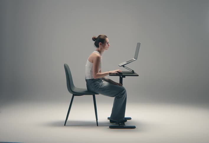 A woman in casual attire sits on a chair in a bright studio working on a laptop elevated by a stand on a small table demonstrating proper ergonomic posture in a side profile view