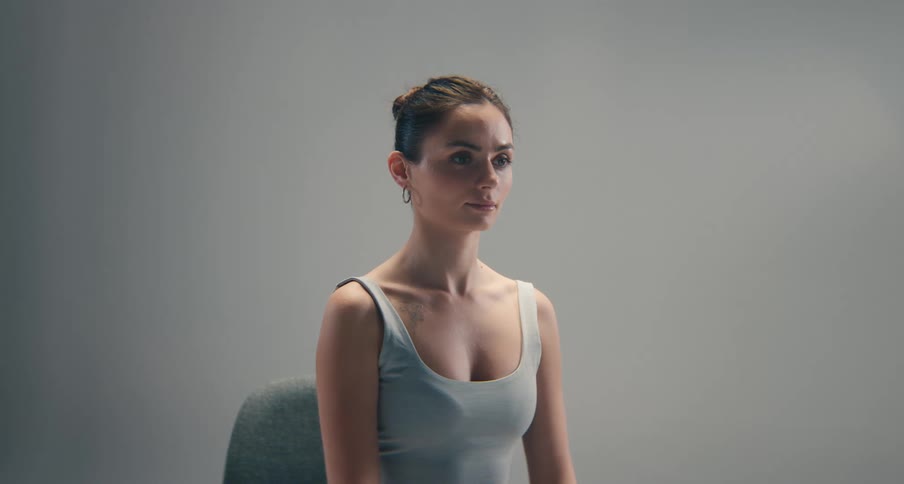 A closeup shot shows a woman wearing a gray tank top seated with a neutral expression as she looks slightly offcamera in a bright minimalist studio setting