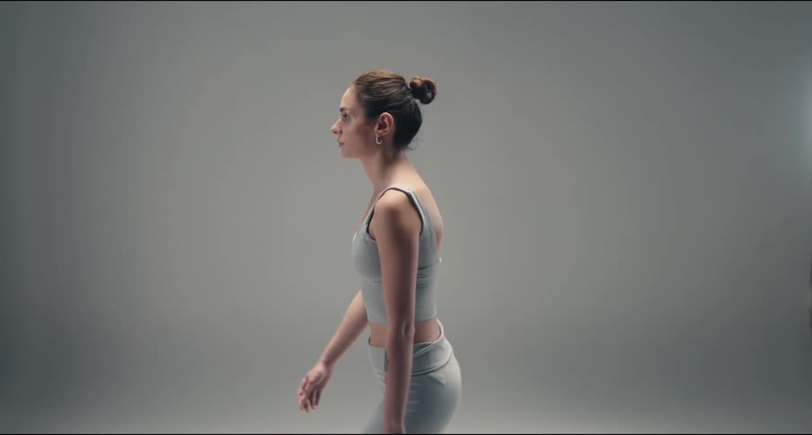A focused woman in athletic wear stand and walks in profile against a neutral grey background