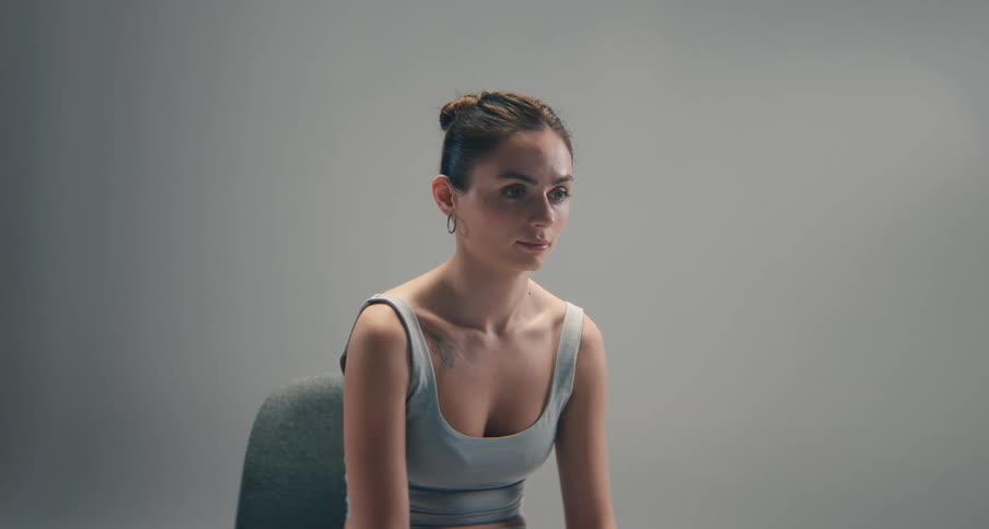 A medium shot of a young woman with her hair in a bun wearing a gray tank top sitting on a chair looking down and away from the camera in a professional studio setting