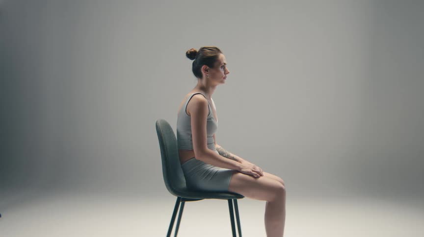 This video shows a static sideprofile shot of a woman seated on a modern chair in a studio setting she is wearing athletic clothing a grey sports bra and shorts and has her hair pulled up in a bunshe maintains a neutral upright posture and gazes