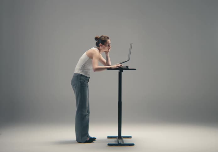 A woman in a white tank top and loose blue jeans is standing at a small high black table leaning forward with her elbows resting on the surface she is looking intently at a laptop placed on the table demonstrating poor posture