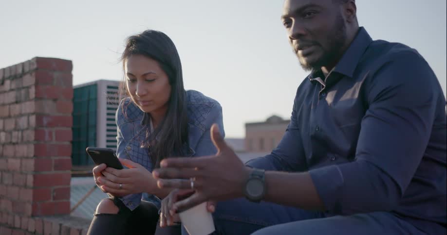 A woman uses a cellphone as a man talks while they are sitting outside