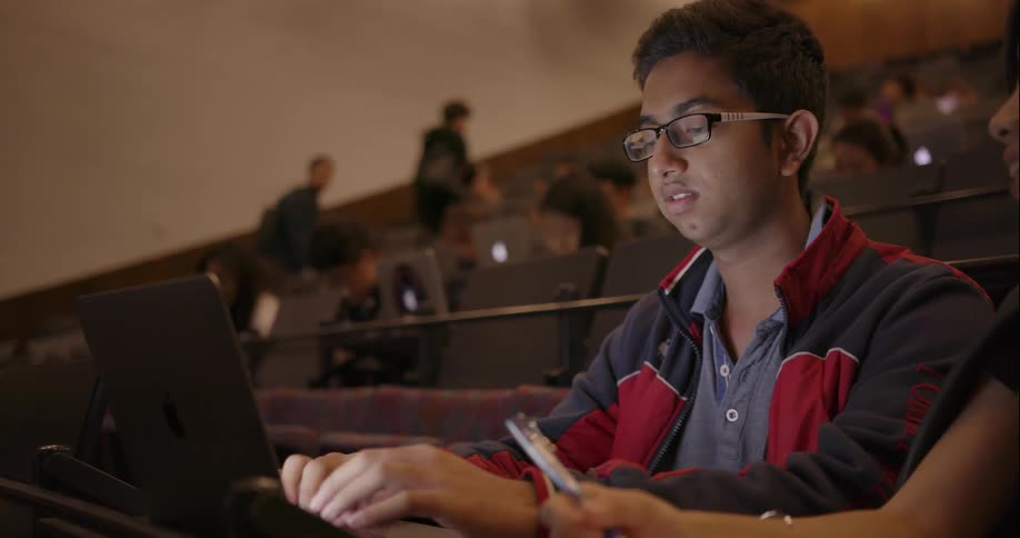 A student sits in a busy auditorium and types on his laptop