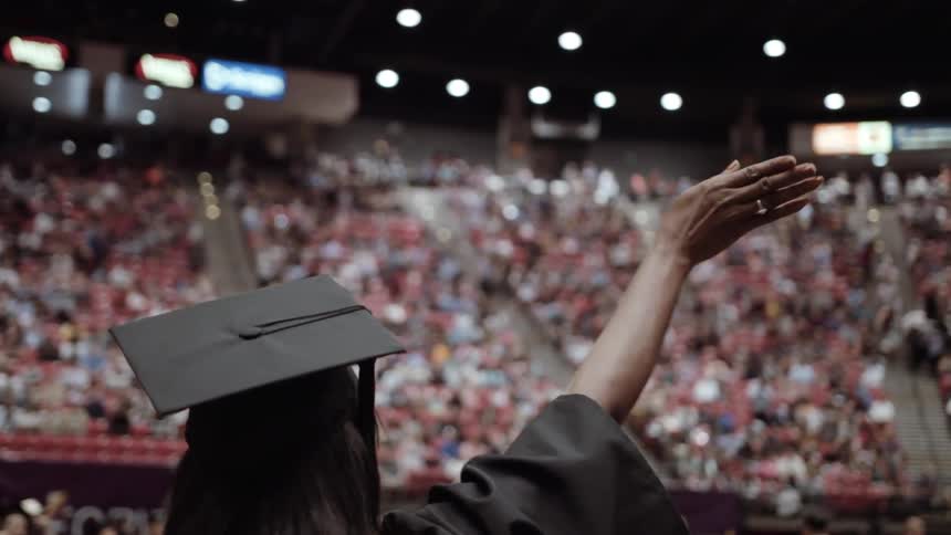 A woman in a cap and gown waves at a graduation ceremony