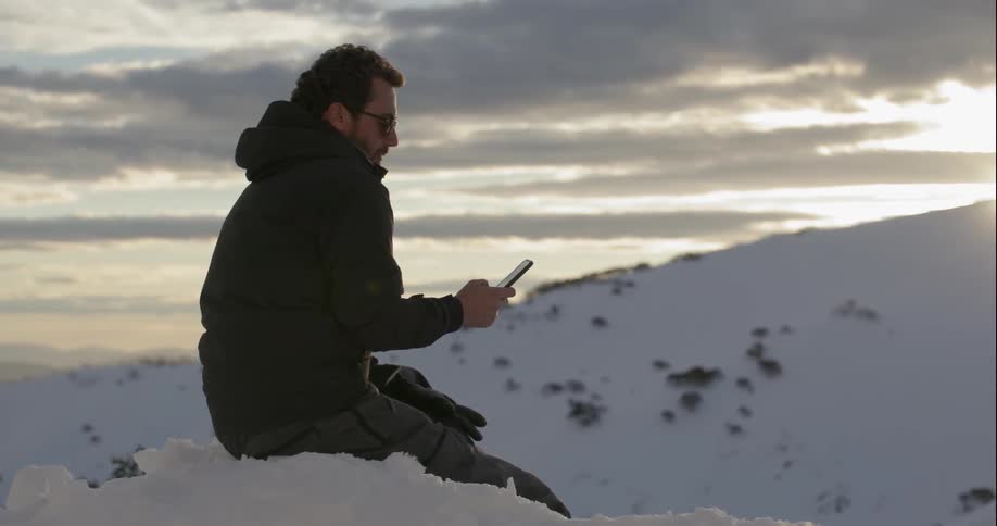 A man sits in the snowy mountains while using his phone