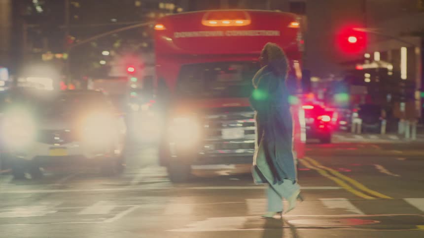 A person wearing a long coat and widelegged pants is crossing a busy city street at night walking through a crosswalkthe scene is dominated by bright car headlights and red traffic lights suggesting a lively urban setting