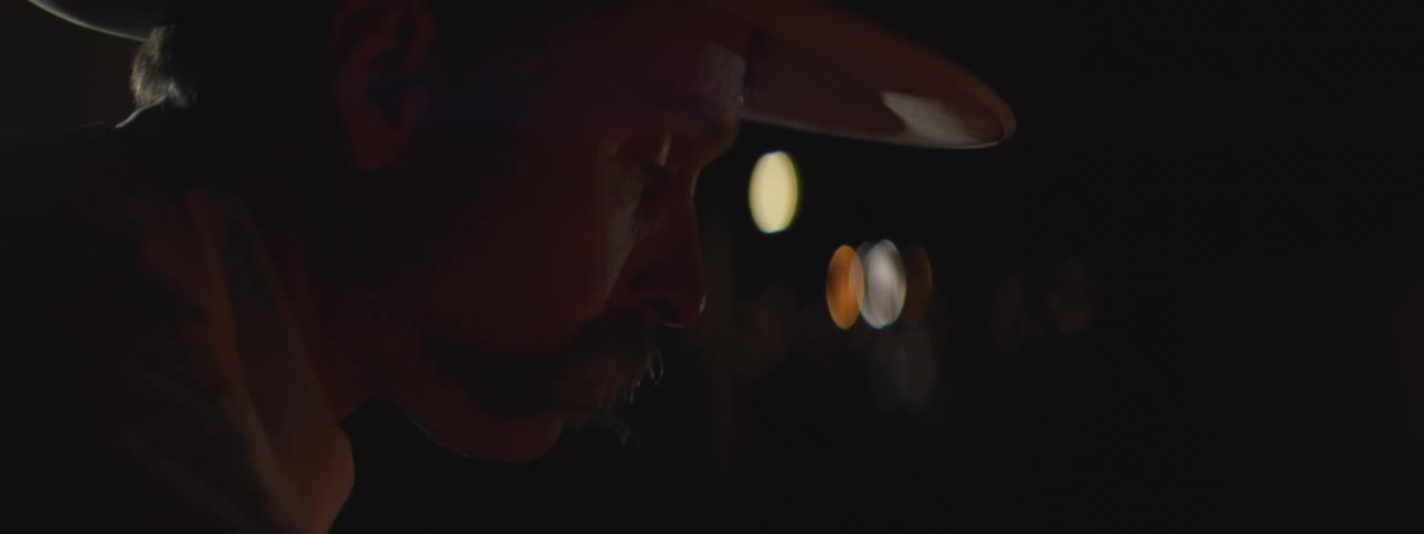 Man wearing a hat leaning forward in a dark setting with blurred lights in the background