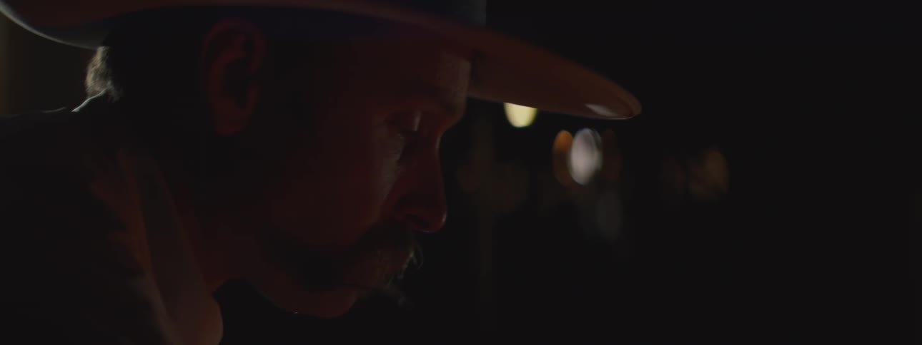 A man wearing a cowboy hat and mustache looks downward in a dimly lit environment