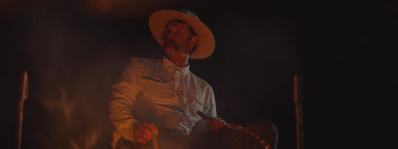 Cowboy sitting near a campfire holds a stick while looking to the side