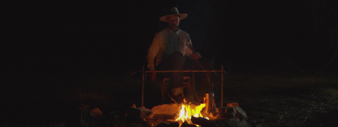 A man wearing a cowboy hat and lightcolored shirt is sitting by a campfire in a dark outdoor settinghe is positioned behind a cooking rack suspended over the flames with a metallic mug nearby