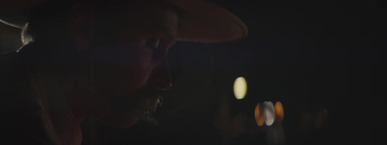 Man wearing a western hat sits outdoors at night outside a building