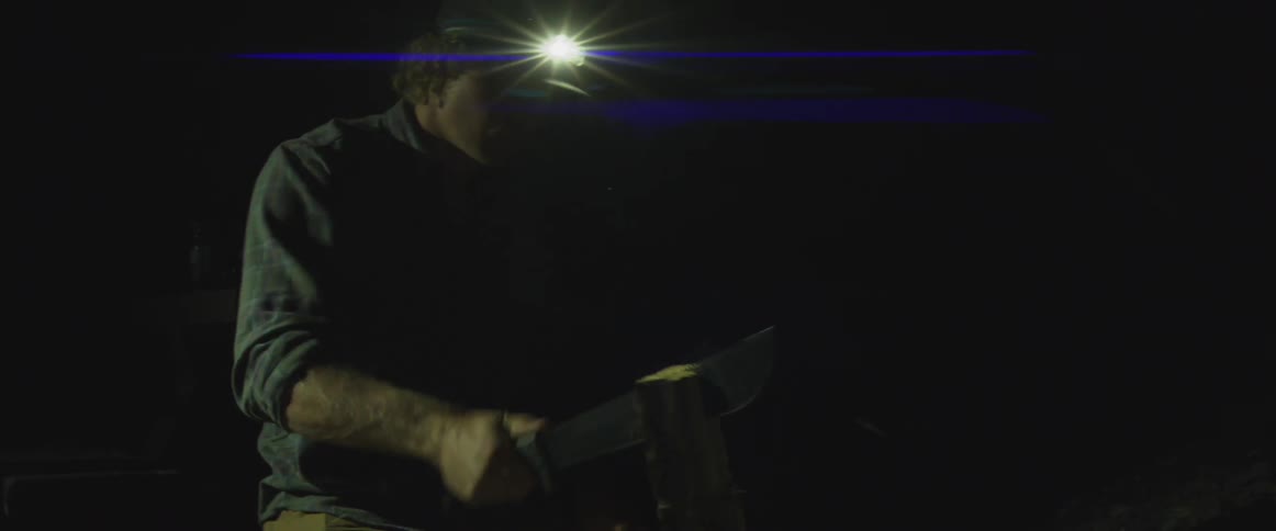 A man wearing a hat with a flashlight in a dark environment using a machete to strike a piece of wood