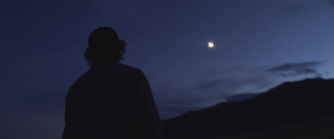 Man standing outside facing mountains as the moon glows in the night sky