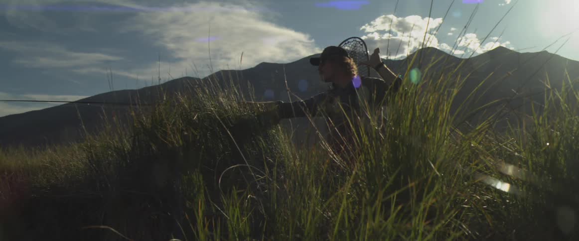 A man stands on a riverbank high in the mountains carefully stretching out his fishing line against the backdrop of rugged nature