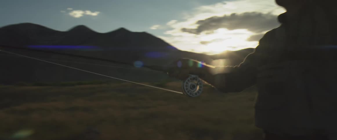 A man backs away from a vehicle while pulling a fly fishing rod out of the trunk against a mountain sunset