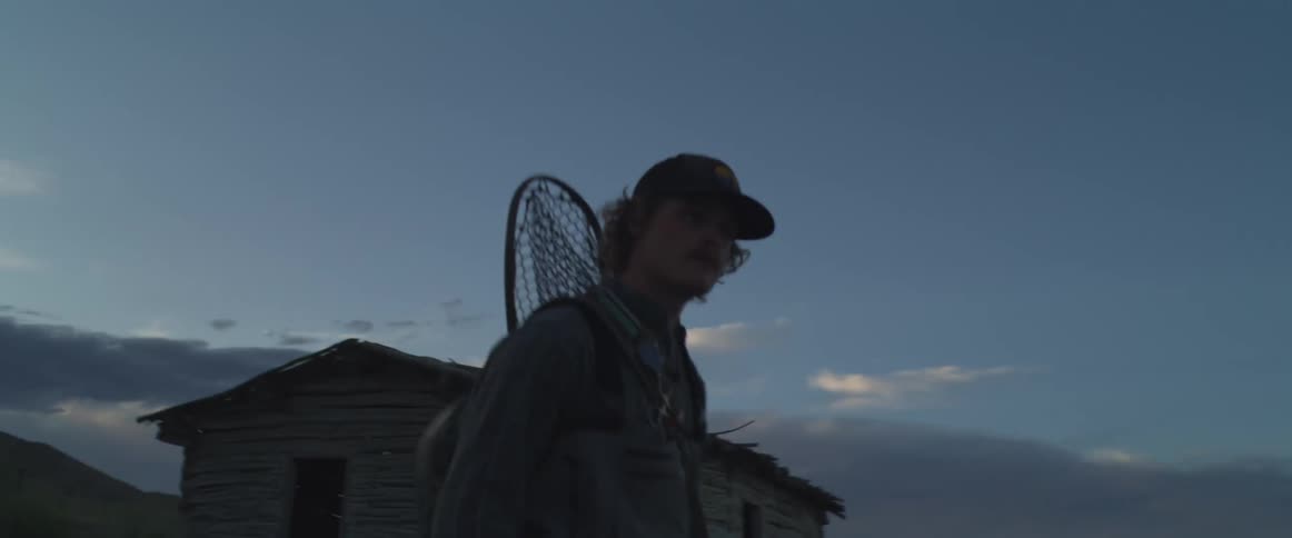 Man walking at dusk holding a fishing rod near a wooden cabin