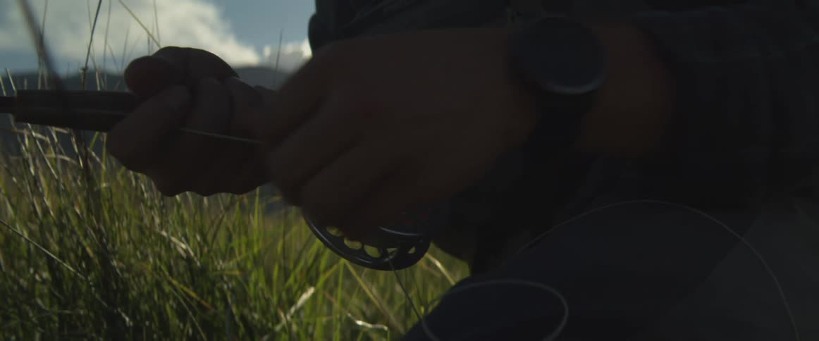 Hands adjusting the reel of a fishing rod amid tall grass