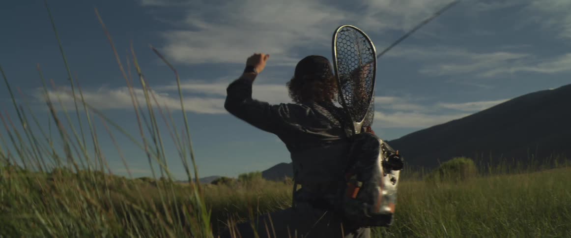 A person wearing a cap a vest and a backpack with a fishing net attached is fly fishing in a tall grassy fieldthey are kneeling slightly and casting their line with a fishing rod against a backdrop of mountains and a blue sky