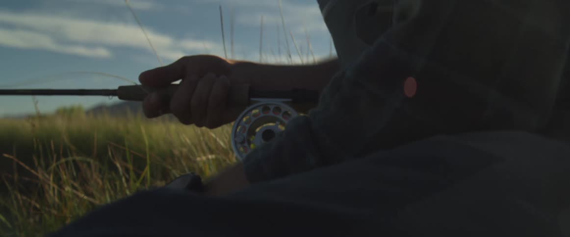 Man fishing crouches in a field of grass pulling fishing line on a cloudy day