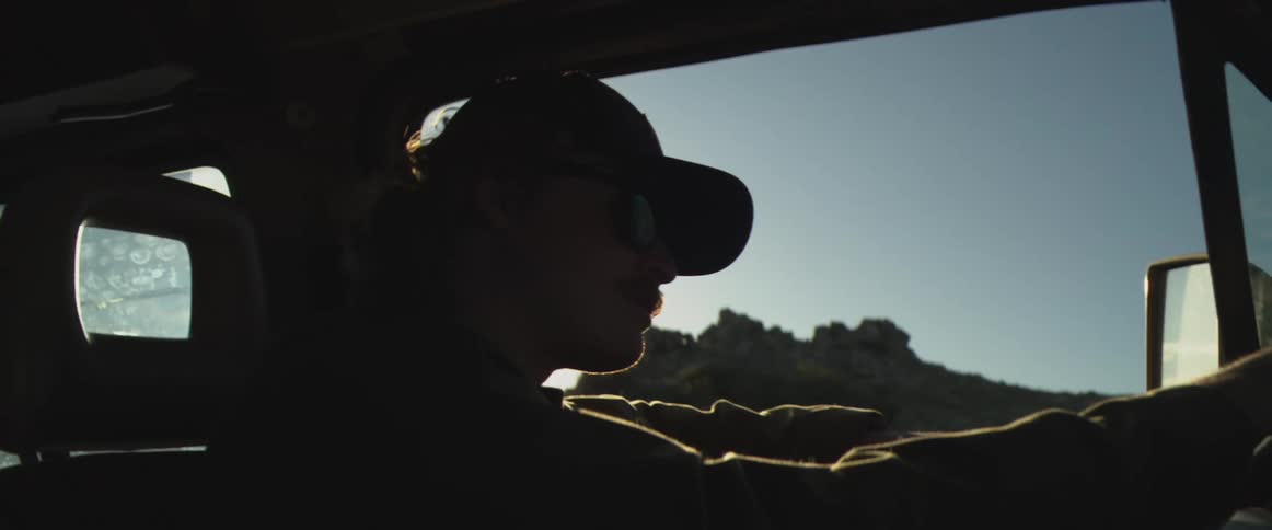 Man wearing a cap driving a vehicle with a sunlit landscape in the background