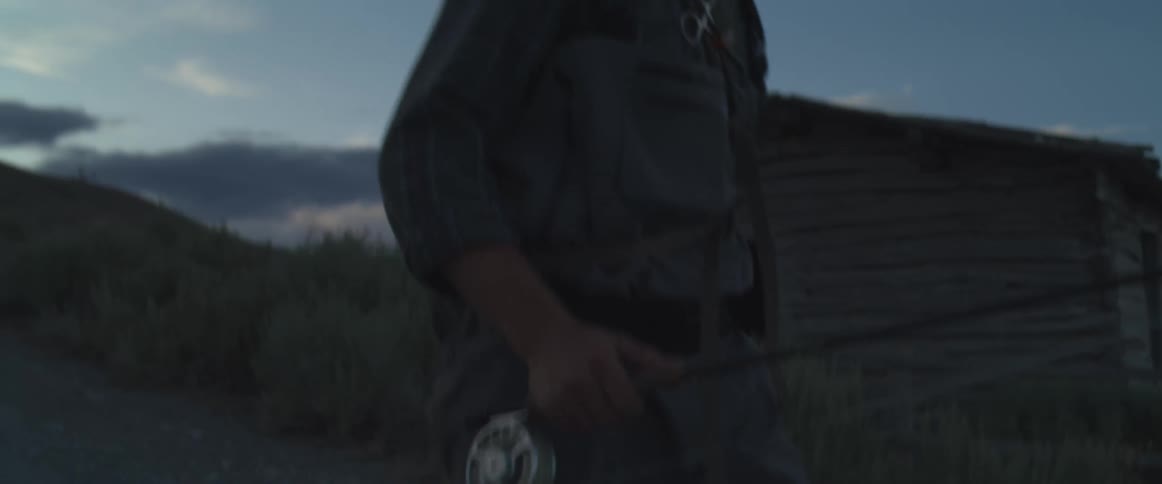 A fisherman carrying a rod and net walks past a rustic cabin at dusk