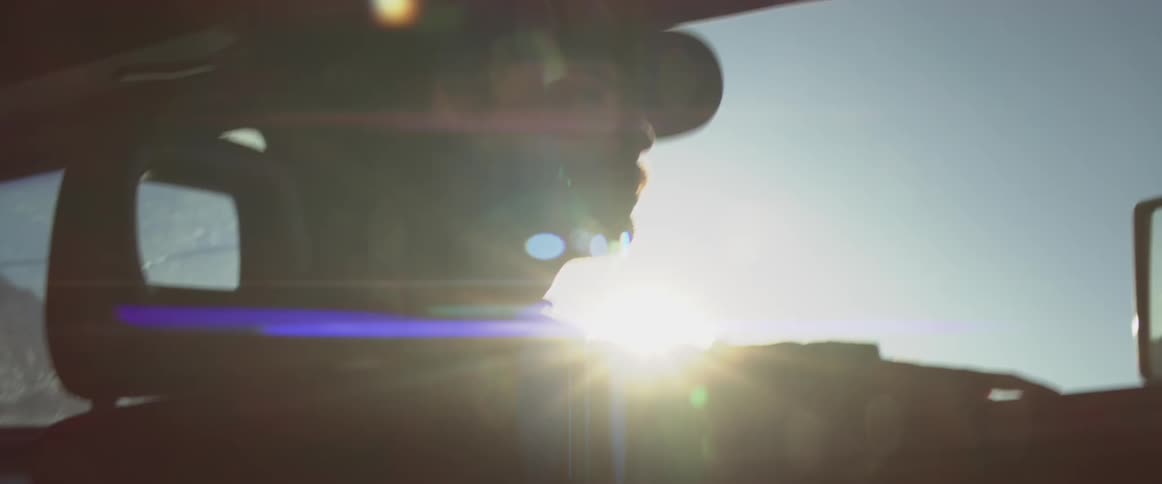 Driver wearing a cap drives with one hand resting on the door as sunlight enters the vehicle