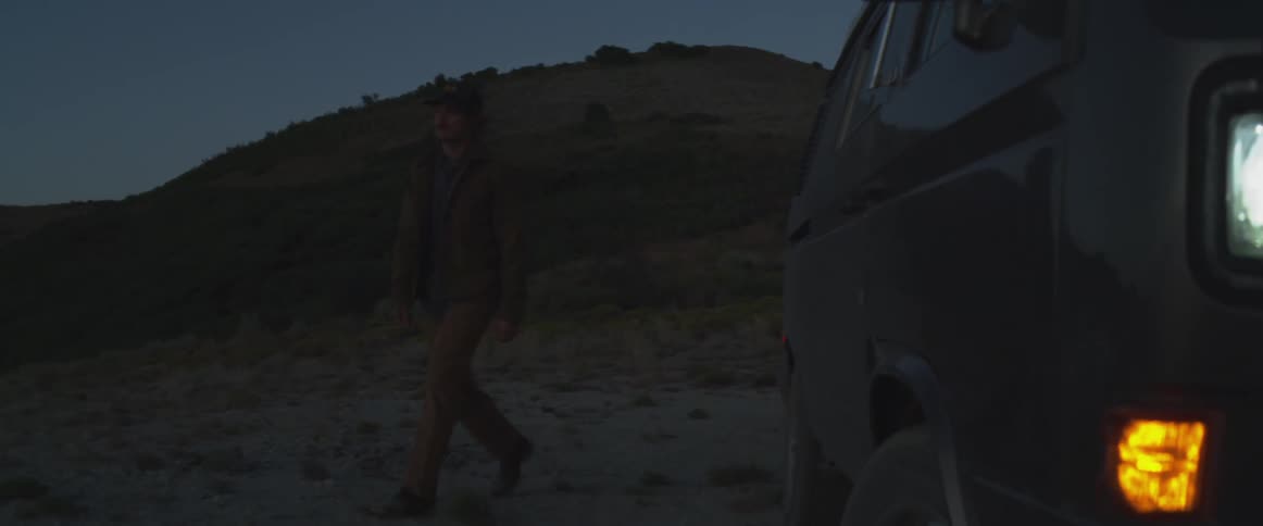 A man wearing a cap is walking in a desert environment and next to him is a car with its headlights on