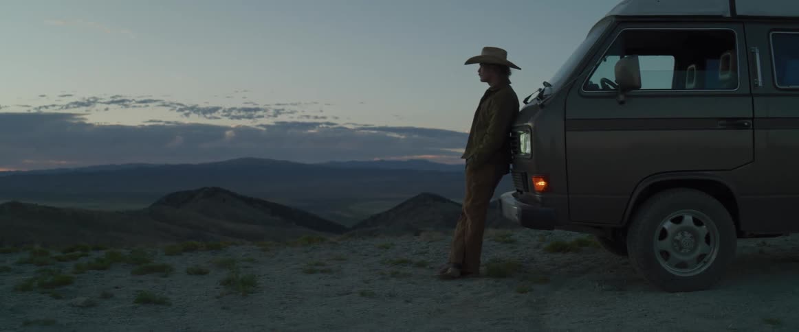This video shows a wide shot of a person standing next to a van at a scenic overlook likely near dusk or dawna person wearing a cowboy hat a jacket and pants is leaning against the front fenderside of a darkcolored van possibly a classic volkswag