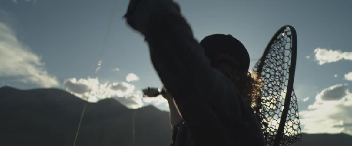 A close view of a man wearing a cap and sunglasses fly fishing and casting his line against a bright sun flare with a net over his back and mountains in the background