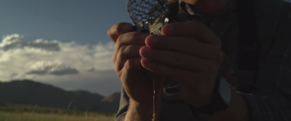 The video shows a closeup backlit shot of a person likely a fly fisherman wearing a hat and sunglasses in a grassy mountainous field at dusk or dawnthe person is intently focused on their hands carefully working with a fishing line and a small lu