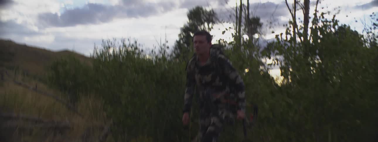 A hunter in camouflage walks through the brush carrying a compound bow