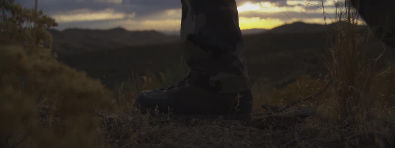 A person wearing a pair of hiking boots walks past a mountain landscape at sunset