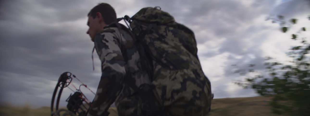 A man in military uniform carrying a backpack and holding a bow walks through bushes while hiking up a mountain on a cloudy day