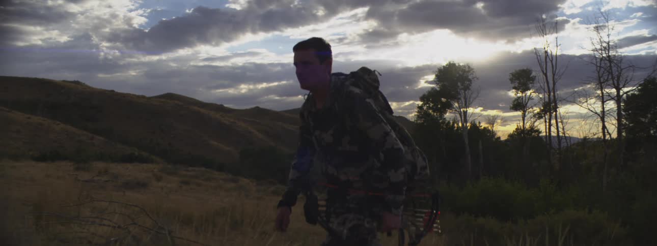 A man hikes up a hill at sunset carrying a backpack and holding a bow as he ascends