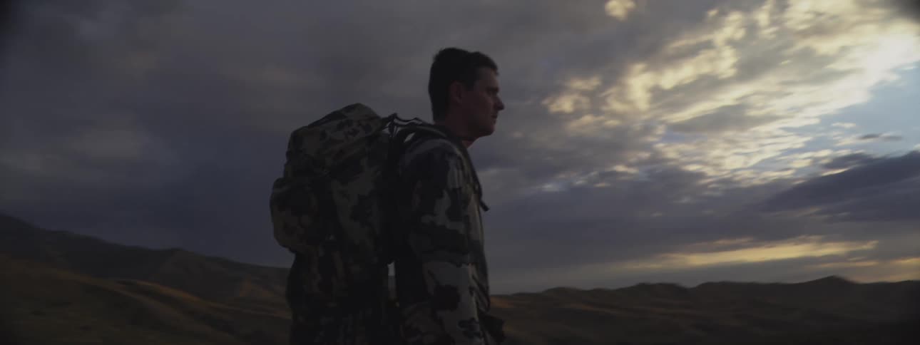 Man wearing a backpack and camouflage clothes stands looking at the sun setting over mountains in the distance