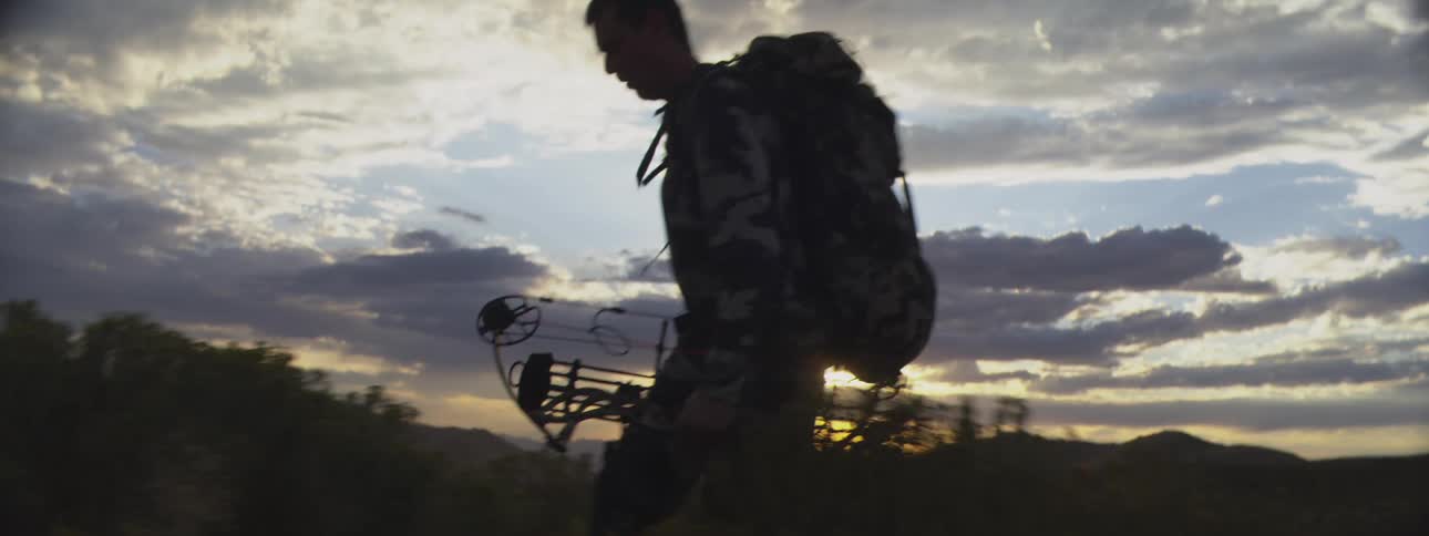 Man in camouflage gear walking with a backpack and a compound bow at sunset
