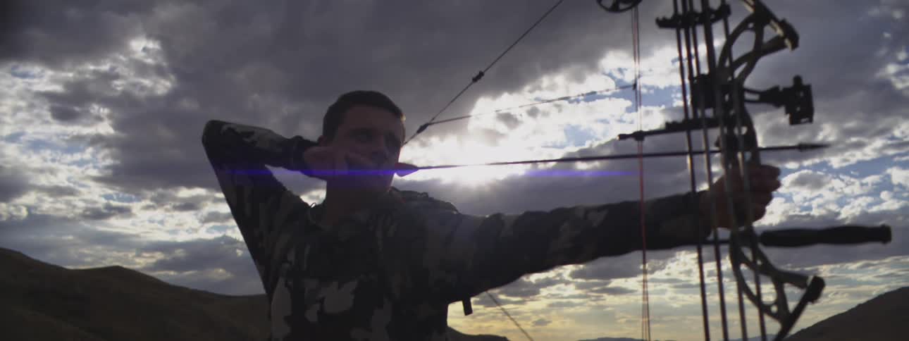 Man stands holding his bow with an arrow pulled back on the string ready to shoot