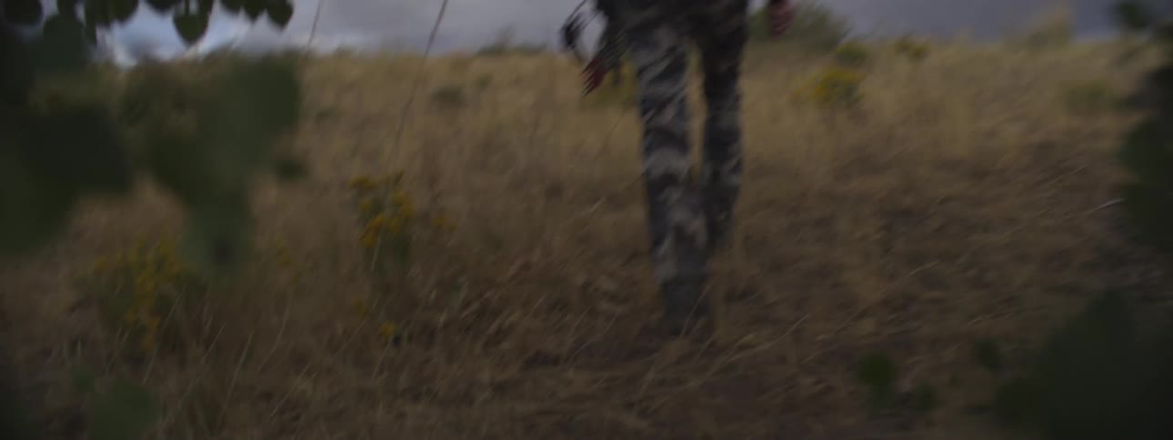 A man hikes up a mountain trail on a cloudy day dressed in a military uniform carrying a backpack and holding a bow