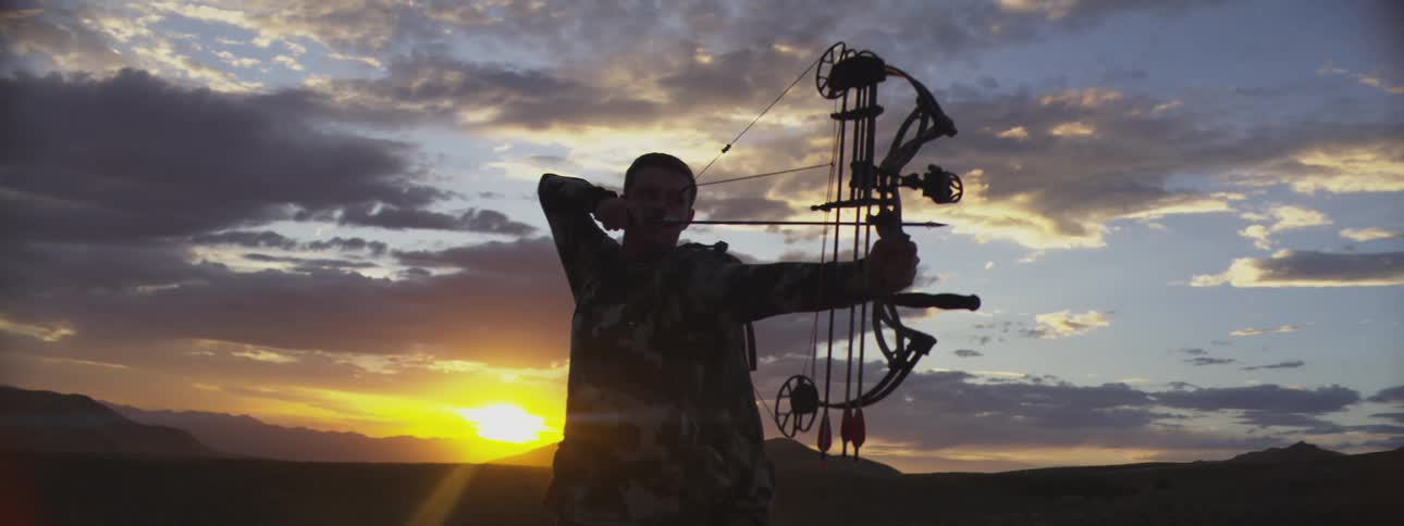 A person in camouflage clothing is seen aiming a compound bow at sunset silhouetted against a dramatic cloudy sky and distant hillsthe archer draws the bowstring back fully and then releases the arrow though the release is cut off in the final frame