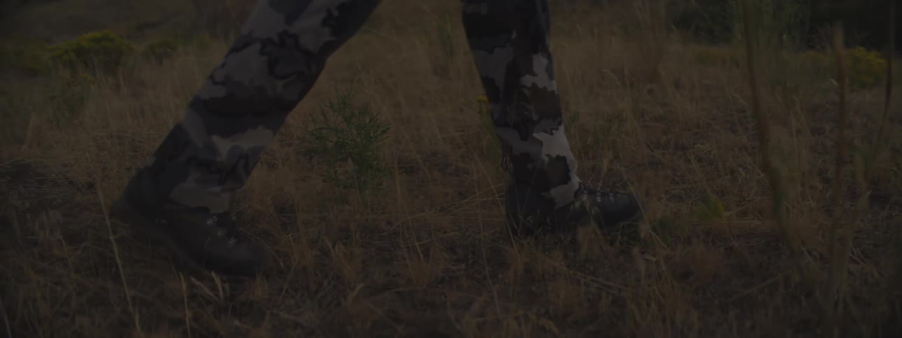 His short video clip shows a persons legs walking through an area of dry grass and lowgrowing plantsthe person is wearing camouflage pants grey black and white pattern and dark hikingutility bootsthe camera focuses on the legs and feet as the