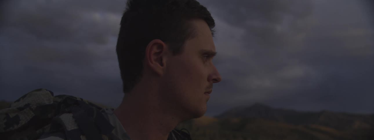 A closeup shot features a mans profile looking offcamera with a serious or focused expressionthe man is outdoors at dusk with dark stormy clouds and mountains visible in the background