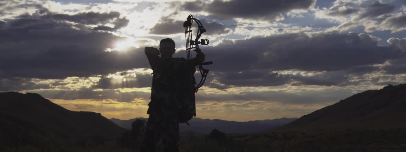 A hunter silhouetted against a dramatic sunset sky is drawing back a compound bowthe person stands on a high point overlooking a valley ready to shoot
