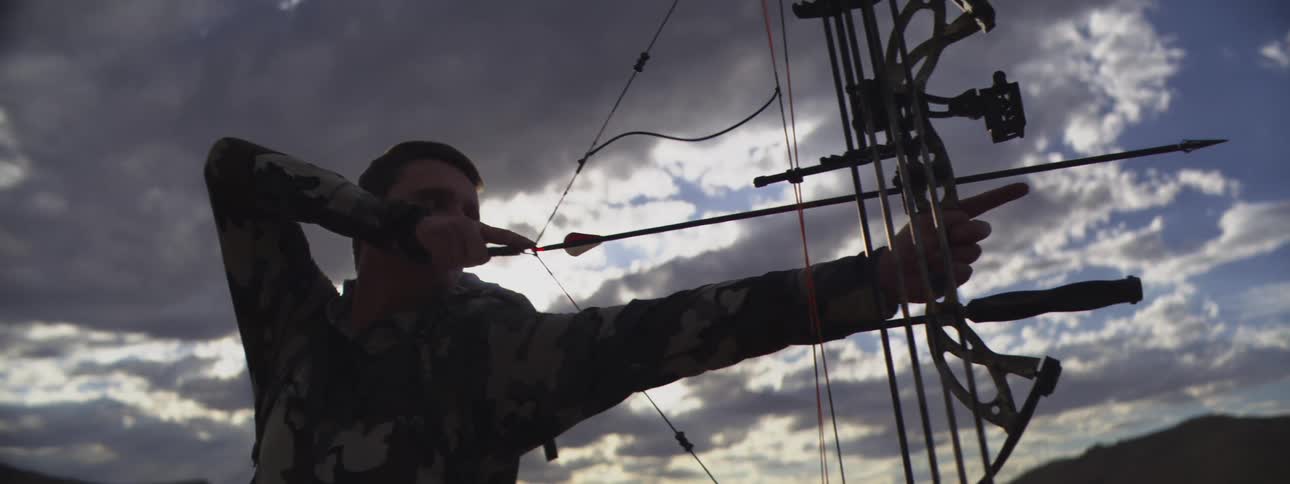 Man in camouflage gear drawing a compound bow with an arrow aimed