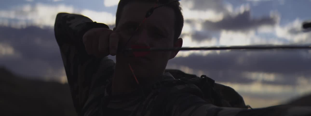 A man is seen in a closeup shot heavily silhouetted against a bright cloudy sky at dusk or dawnhe is focused and drawing back an arrow on a compound bow suggesting he is bow hunting or target shooting outdoors