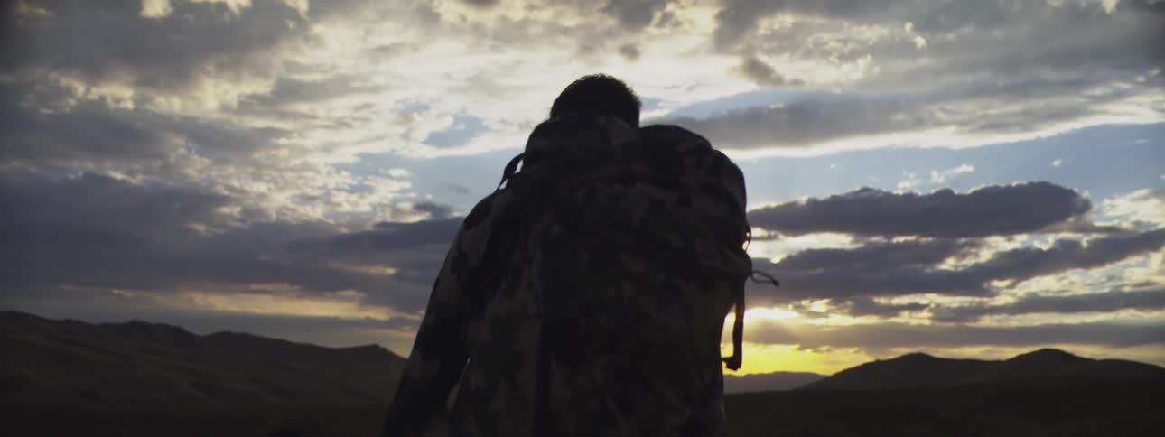 A man hikes through the wilderness at sunset carrying a large backpack and pausing to observe his surroundings