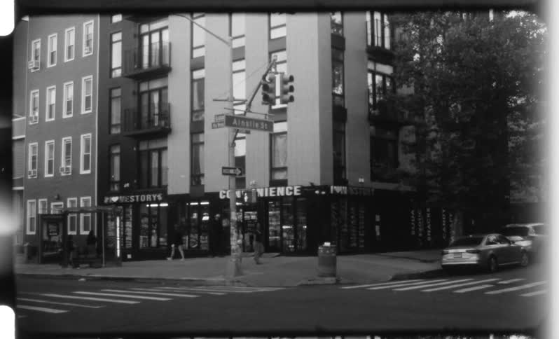 A black and white still shot captures a wide view of an urban corner showing multistory apartment buildings a convenience store and a street sign for ainslie st with people walking and cars parked nearby